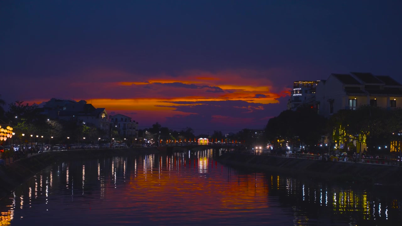Magnifiques nuages de coucher de soleil orange ardent se reflétant dans la paisible rivière Hoai au crépuscule dans la vieille ville de Hoi An au Vietnam, avec des bâtiments riverains éclairés par des lanternes et un pont.