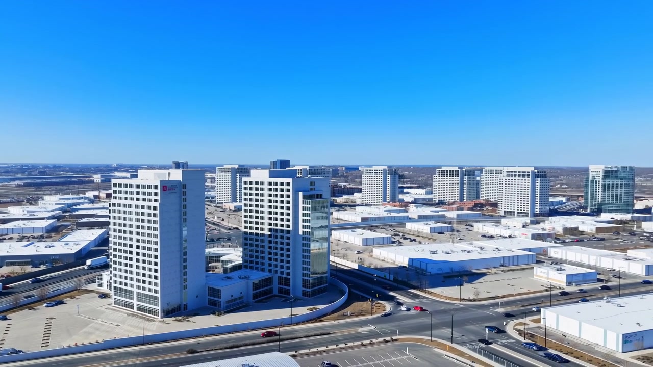 Vista aérea de gran angular que muestra tres torres blancas de gran altura conectadas con detalles de esquina de vidrio y fachadas de ventanas en cuadrícula, situadas junto a rampas de autopista curvas, una rotonda, vehículos en movimiento y un espacio urbano abierto.