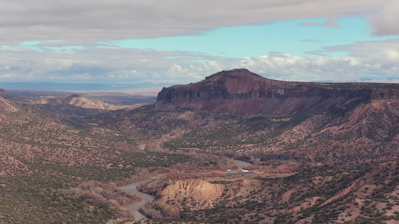 A slow lateral move captures the textured canyon cliffs and winding Rio Grande below, highlighting layered rock formations and high-desert terrain