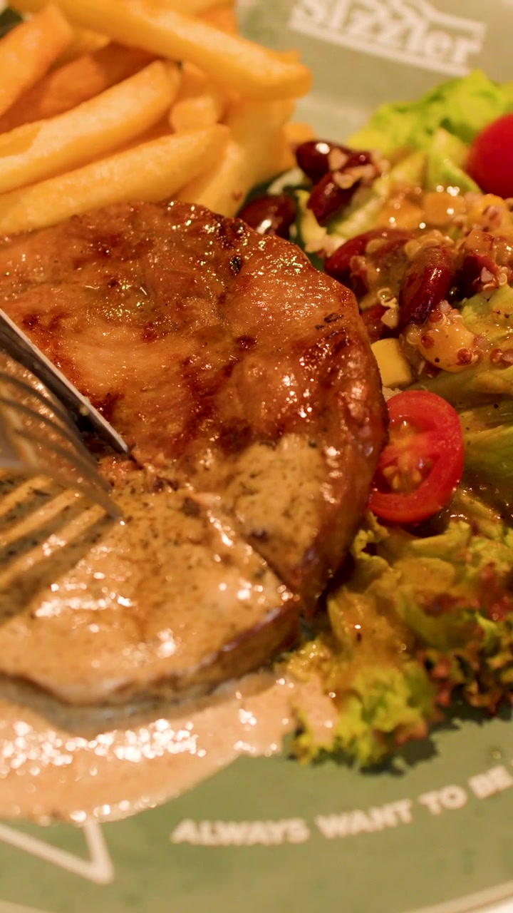 Una persona usa un coltello e una forchetta per tagliare una bistecca di maiale alla griglia con insalata mista.