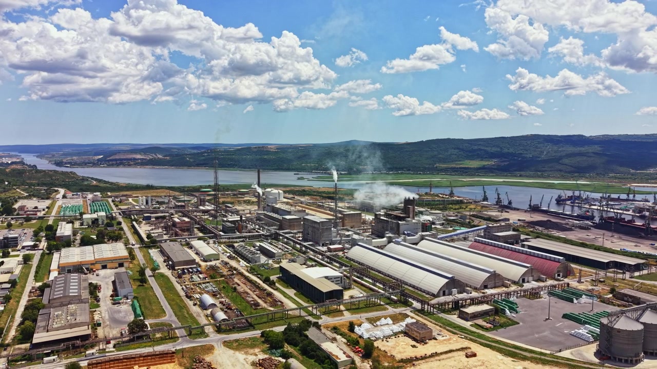 Industrial facility located by a river with buildings and smoke stacks on a sunny day surrounded by hills and clouds