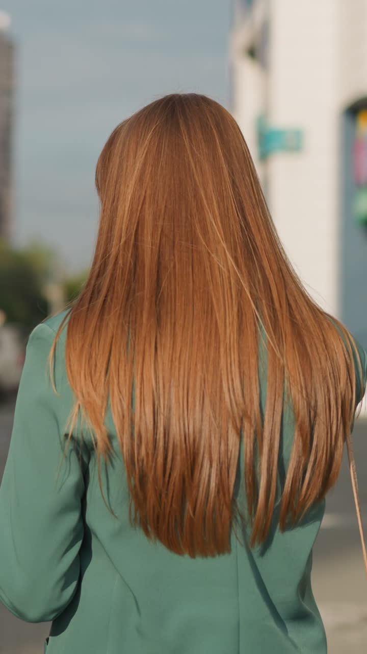 Restless young female with straight long hair talks on mobile phone. Person moves along sidewalk past parked cars on blurred background close backside view