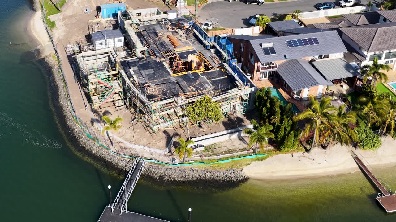 Aerial footage shows a modern house under construction beside a canal, with adjacent homes, palm trees, and clear daylight. Smooth camera movement reveals site details