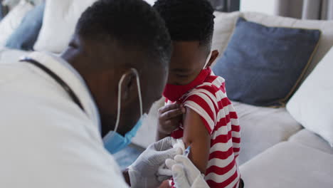 Médico-Afroamericano-Con-Mascarilla-Inyectando-La-Vacuna-Covid-19-A-Un-Niño-En-Casa