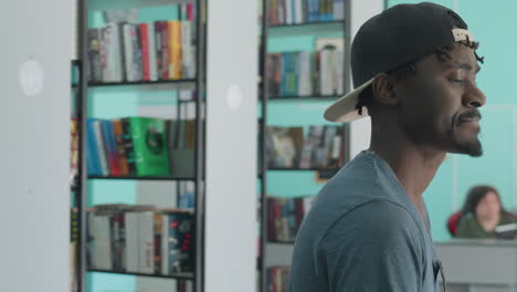 close up side view of young man wearing cap and casual shirt standing among bookshelves in bright library interior, looking around with thoughtful expression while surrounded by colorful books