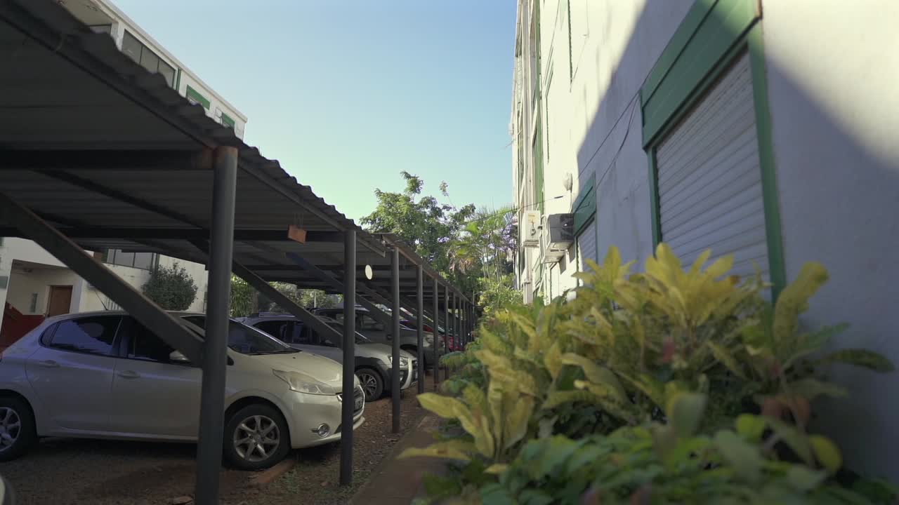 Slow moving camera footage showing apartment building parking lot during sunny afternoon, with vegetation near wall on right and cars aligned on left.