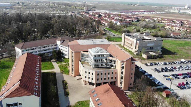 Imagens aéreas de drone sobre o Campus da Universidade de Oradea, com biblioteca, estacionamento e um grande edifício com estudantes.