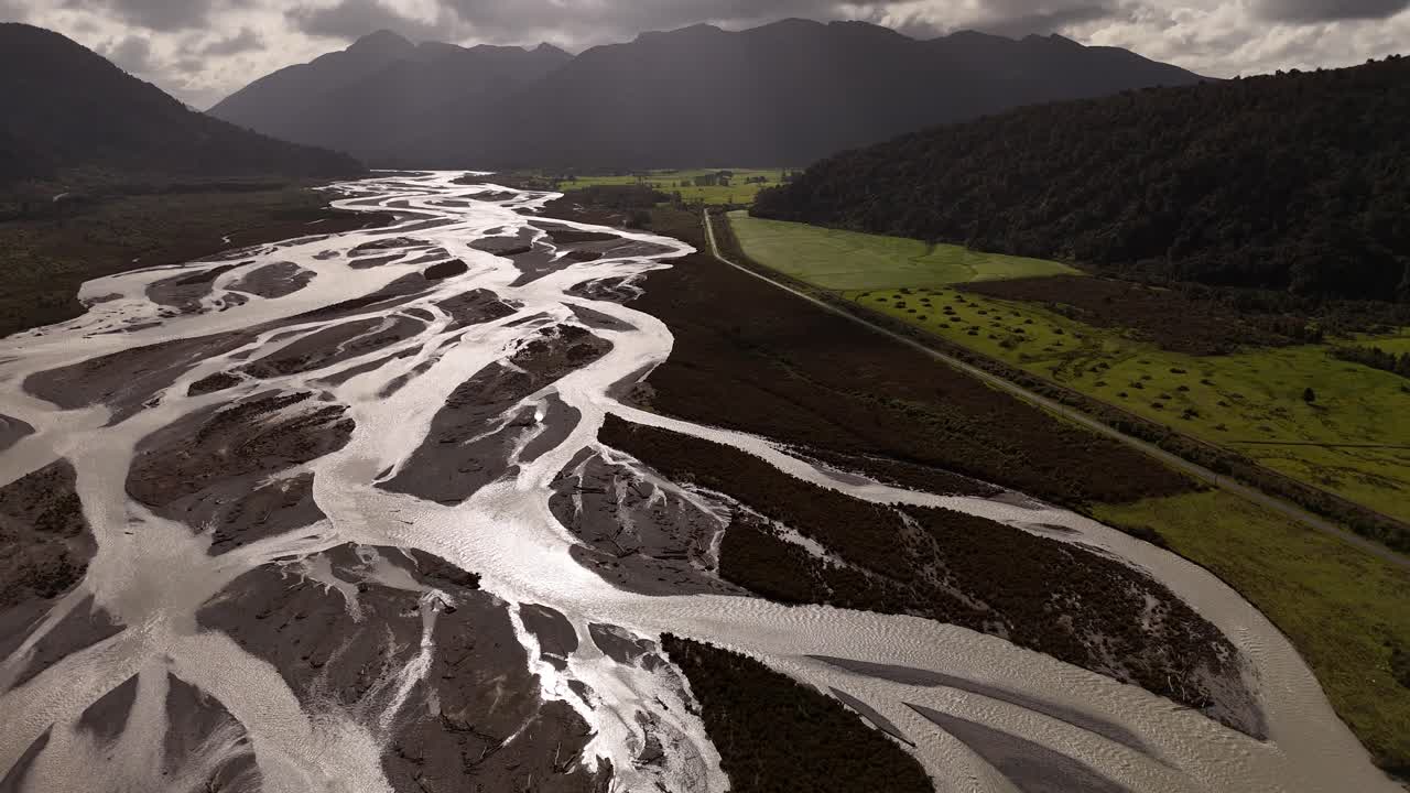 Vista aerea con drone del fiume Bealey a canali intrecciati che scorre attraverso un'ampia valle alpina vicino ad Arthur's Pass sull'Isola del Sud della Nuova Zelanda, circondato da verdi colline, montagne e nuvole spettacolari.