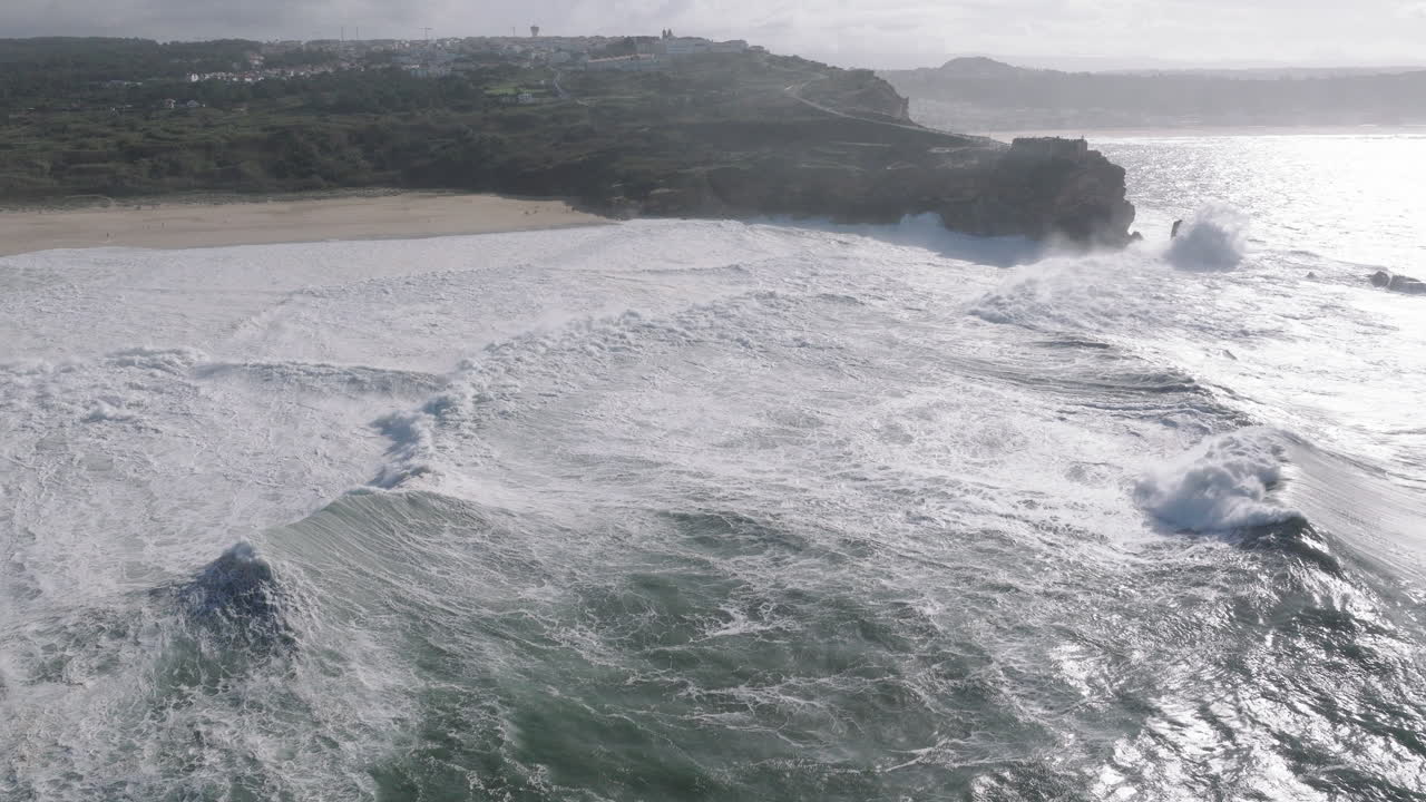 Wide aerial drone shot of big waves coming into shore on a day with giant waves in Nazaré, Portugal, Europe. Flying towards beach and people watching. Big wave surfing town, biggest waves in the world