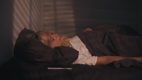 a blonde girl in a white t-shirt sleeps in a gray bed under the rays of the sun in the early morning