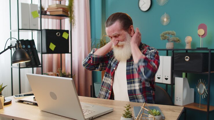 Senior elderly man suffers from spine neck muscle pain at home office workplace working on laptop