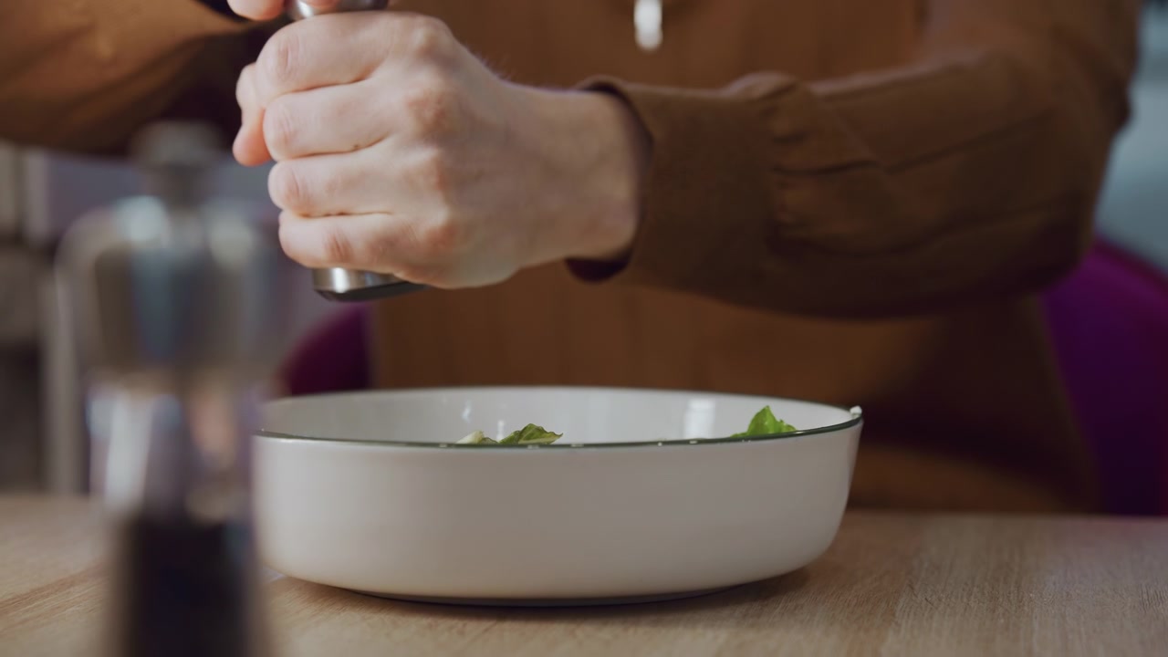 Primo piano di una persona che condisce foglie di insalata verde in una ciotola bianca su un tavolo di legno. Una mano tiene un macinapepe spargendo sale su erbe fresche e verdure. L'illuminazione soffusa esalta la consistenza.