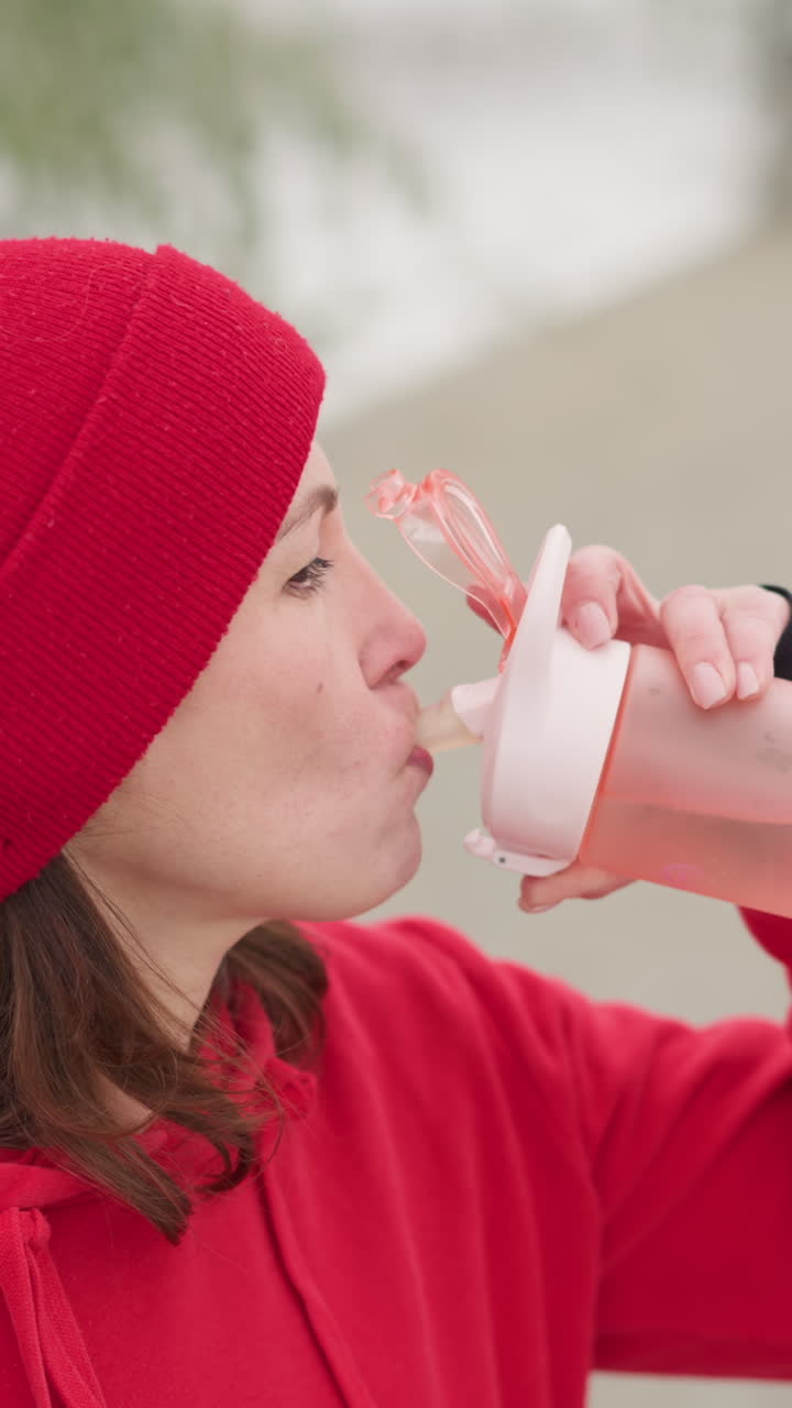 woman in red beanie and hoodie holding pink water bottle outdoors, preparing to drink, surrounded by snowy scenery and blurred bench, set in serene winter setting