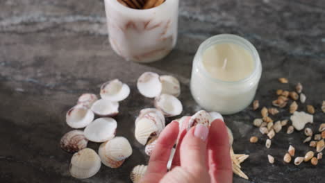 aerial view of person picking up seashell from marble surface observes it then drops it causing other seashells to scatter around decorative candle jar and incense holder in cozy home decor setting