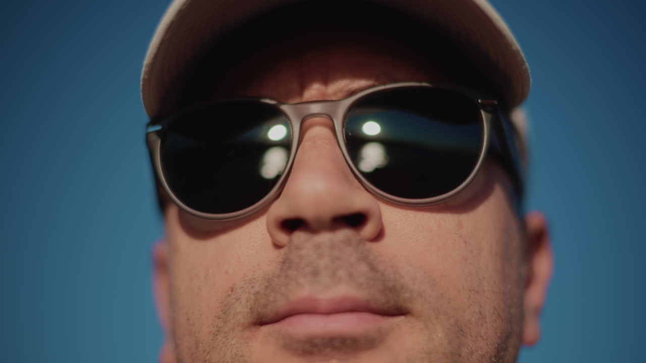 Extreme close up of a man wearing dark sunglasses and a cap, framed tightly against a clear blue sky in strong daylight
