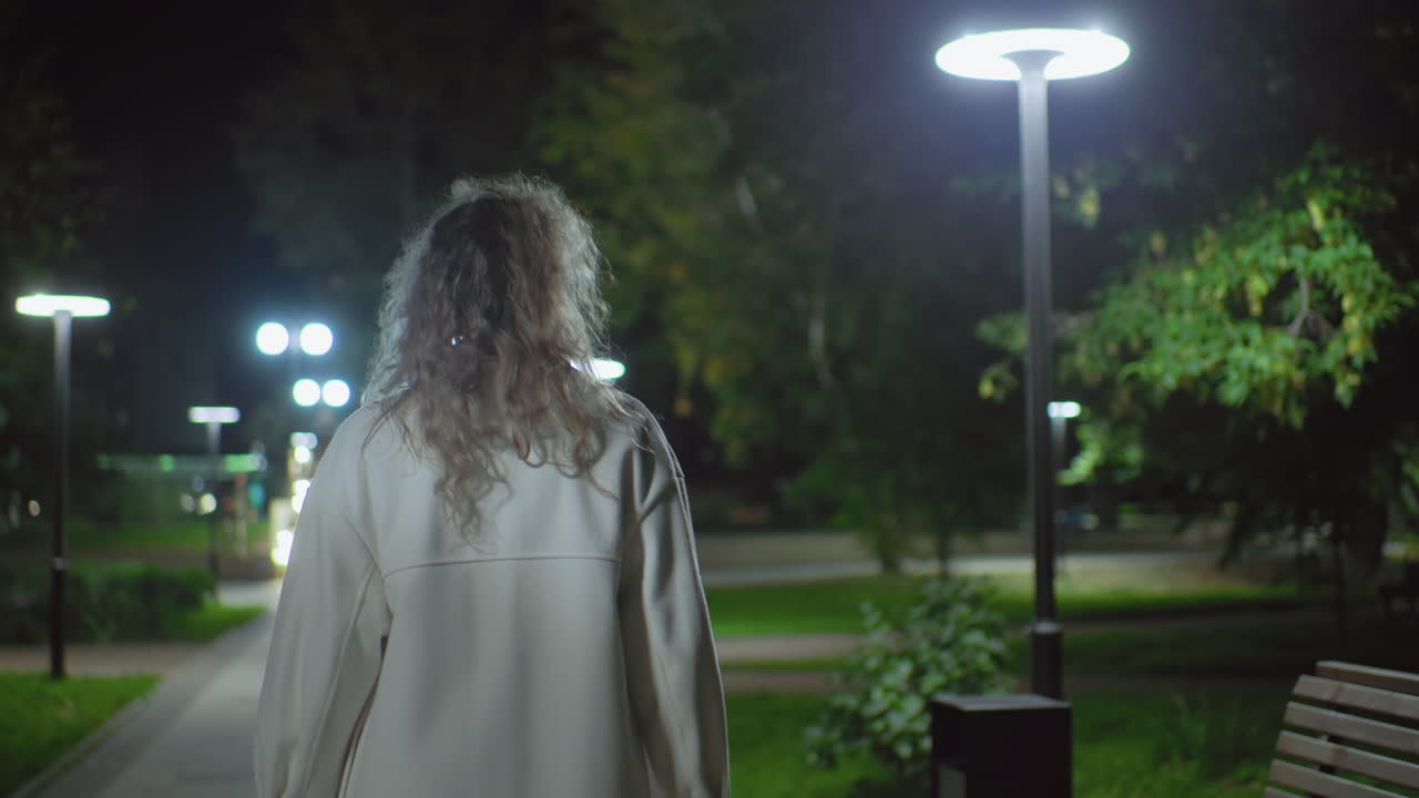 Rear view lady in white coat walking in open park at night, people faintly visible, overhead lamps illuminate surroundings, curly hair gently lit, capturing calm city ambiance with foliage