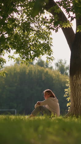 woman sits under large tree with sunlight gently illuminating her, surrounded by lush greenery, background shows distant trees, a football goalpost, and fluttering leaves