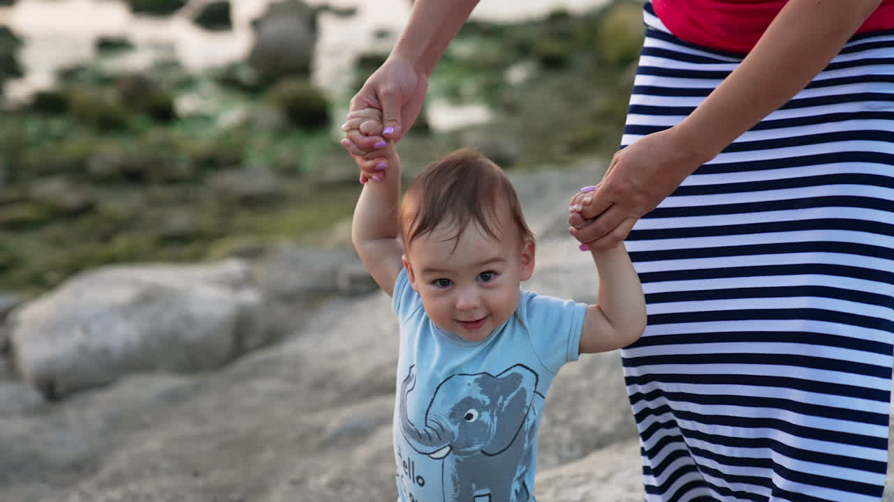 Beautiful Caucasian child walking smiling happily. Mother's hands supporting her baby boy. Nature backdrop in blur.