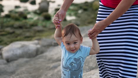 beautiful caucasian child walking smiling happily. mother's hands supporting her baby boy. nature backdrop in blur.