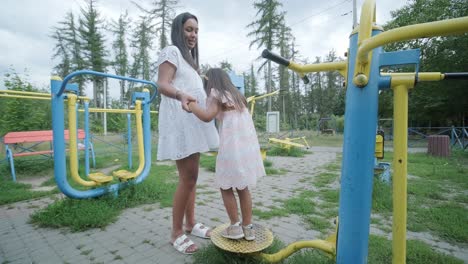 a pregnant mother and her young daughter enjoy playful time together at a playground in the park, surrounded by trees and greenery