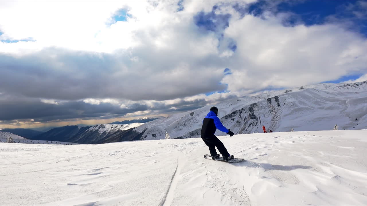Following a snowboarder descending quickly by the slope of the mountain. Fantastic rocky scenery under cloudy sky at backdrop.