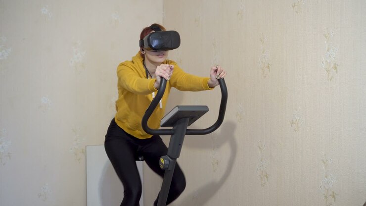A young girl sits and pedals on an exercise bike at home in virtual reality glasses. A girl in yellow sportswear. Home sports training. Close-up. 4K.