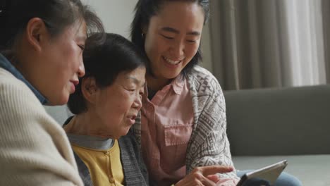 happy senior asian mother, adult daughter and granddaughter in living room using tablet and laughing