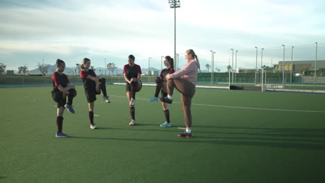 female hockey team warming up with coach on outdoor field, preparing for practice