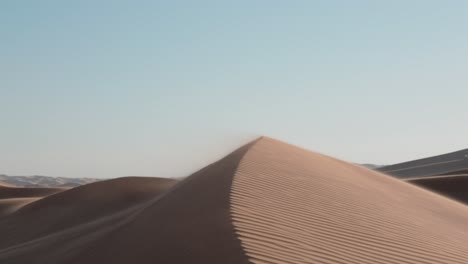 Sand-dune-sharp-edge-in-shadow-as-sunlight-casts-light-on-ripples-and-wind-blows-grains-off-under-blue-sky