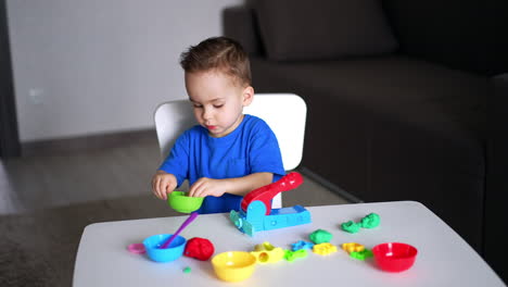sweet baby boy sitting at desk plays with plasticine. toddler takes a piece of plasticine and a stick to cut it.