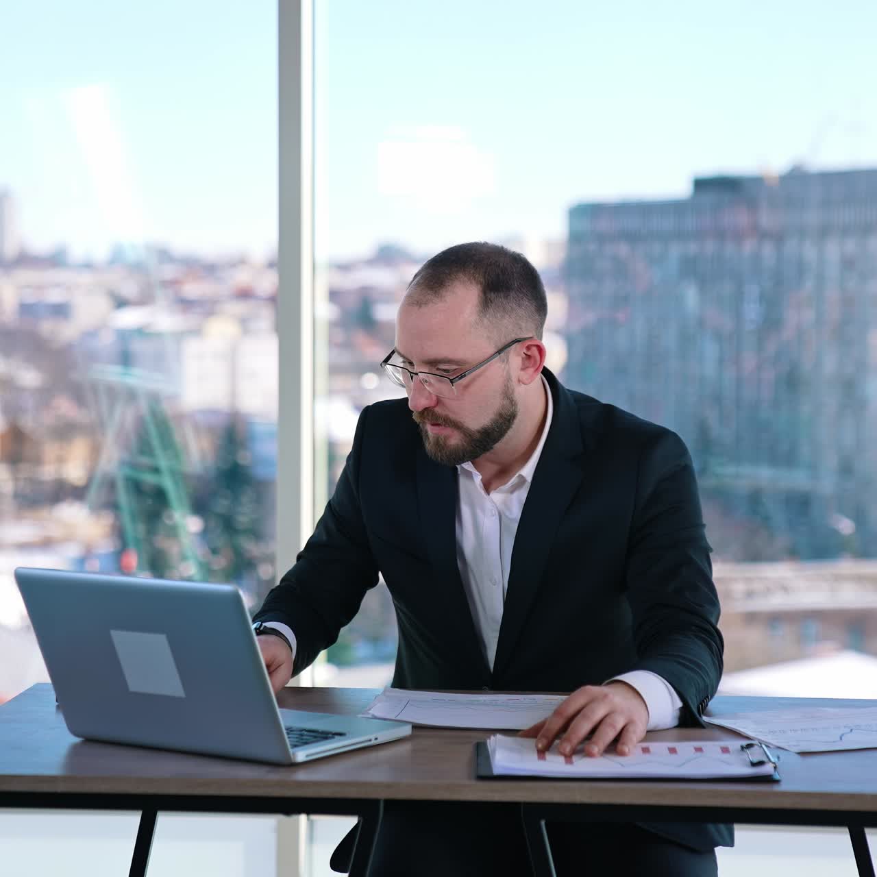 Busy man in suit working in office. Middle-aged entrepreneur in glasses sitting at table with many papers and a laptop and working seriously