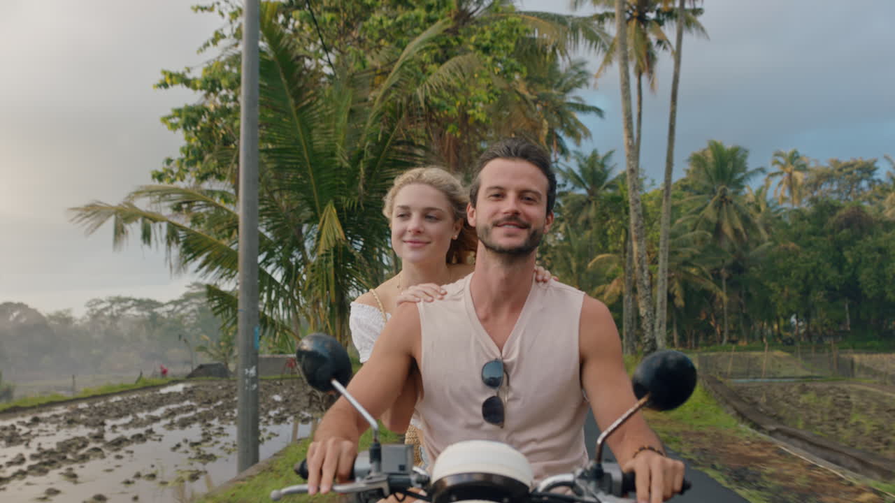 열대 섬에서 스쿠터를 타는 행복한 커플 행복한 여성은 팔을 들고 축하하고 아침 안개에서 오토바이 타고 남자친구와  ⁇ 만적인 모험을 즐기고 있습니다.