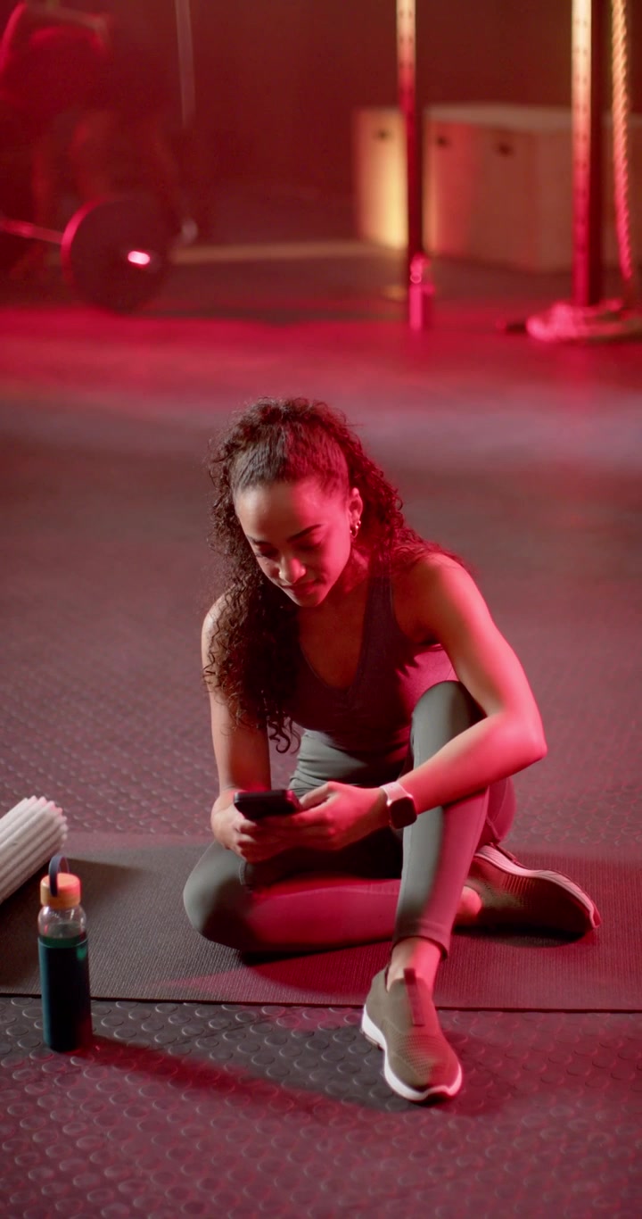 Video vertikal: Wanita Afrika duduk di matras di gym, menggulir ponsel saat istirahat, foam roller di kiri