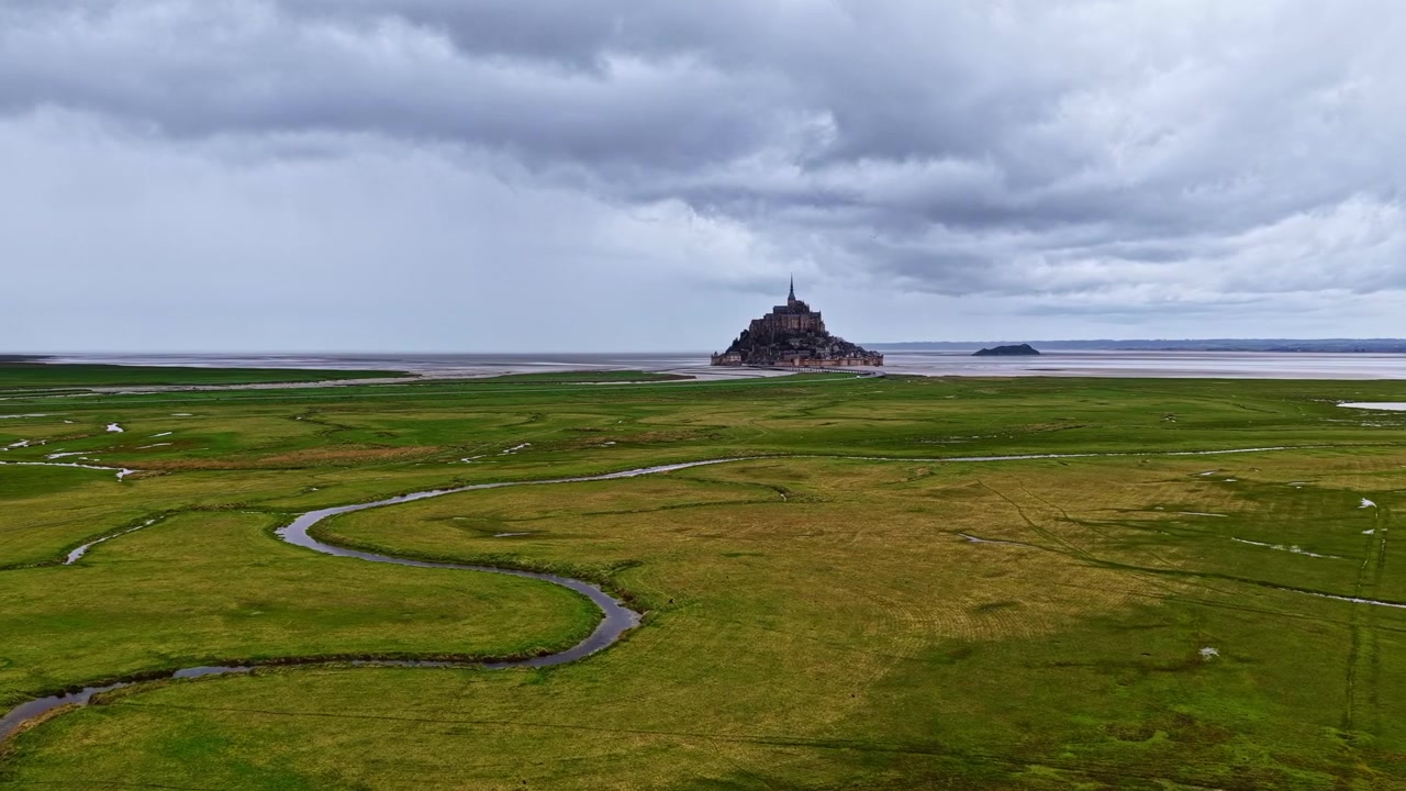 Strömmande vattendrag slingrar sig genom gröna våtmarker mot Mont Saint-Michel, mot en dramatisk molnig himmel. Perfekt för researtiklar eller historiska dokumentärer