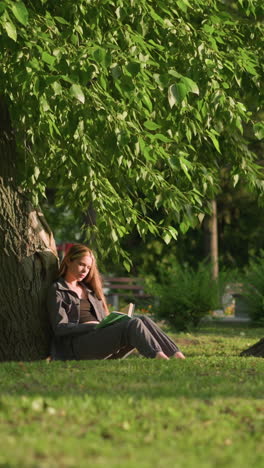 lady sitting outdoors, leaning against tree on grassy field, reading book under, tree leaves swaying gently in breeze, background shows building and people walking slightly blurred in distance