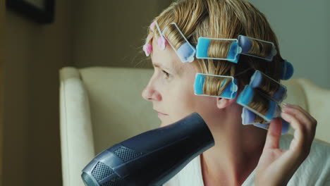 a young woman dries hair with a hairdryer curlers on her head to give shape to her hairstyle