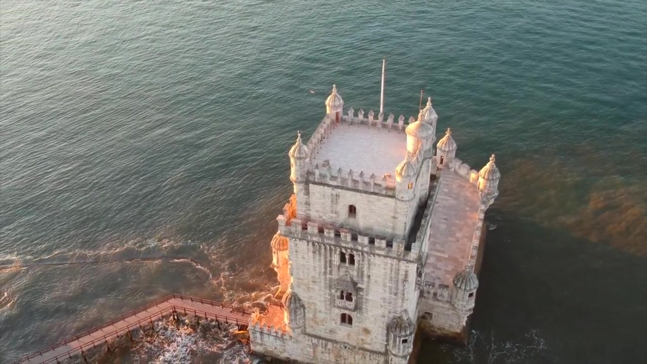 Východ slunce nad lisabonskou pobřežní věží Belém, orámovaný zářící oblohou a klidnými středomořskými vodami, dron stoupá a sklání se dolů.