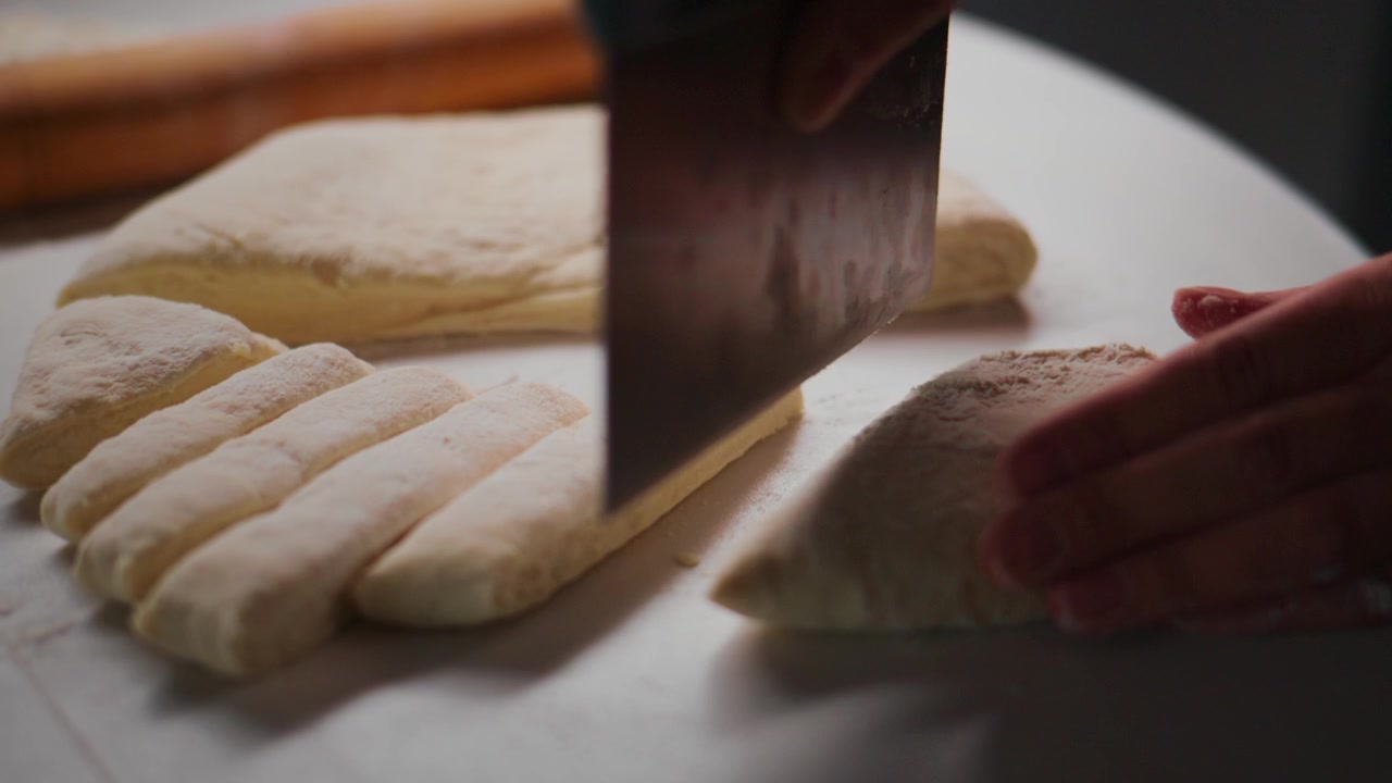 Hand slices fresh dough on a white surface. The knife moves steadily through the soft, pale dough. Rolling pin rests in the background. The action shows precise, calm baking preparation