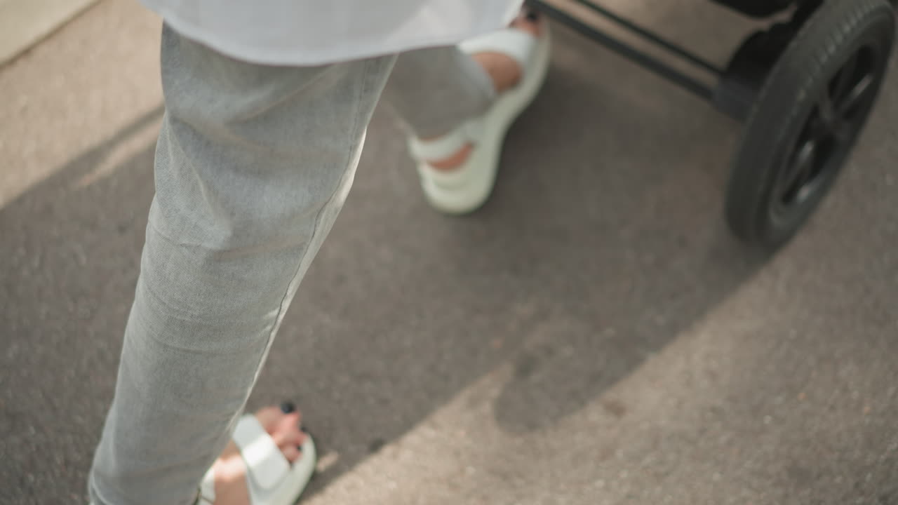 Close-up sandal dan kereta dorong bayi di trotoar; orang tua kota mendorong bayi di kereta dorong saat berjalan di sepanjang trotoar yang disinari matahari; jeans abu-abu dan sandal putih; bayangan roda kereta dorong; momen gaya hidup sehari-hari yang santai