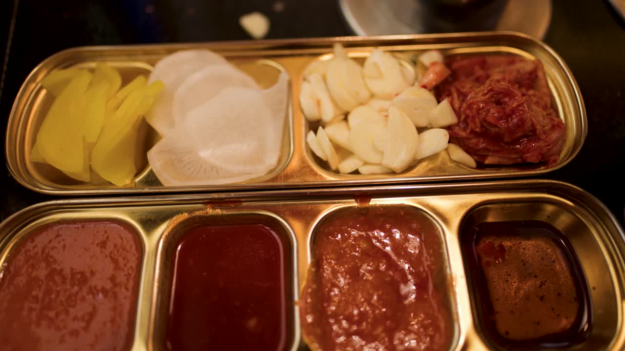 Overhead camera rotates above metal tray with Korean BBQ banchan, garlic, kimchi, dipping sauces