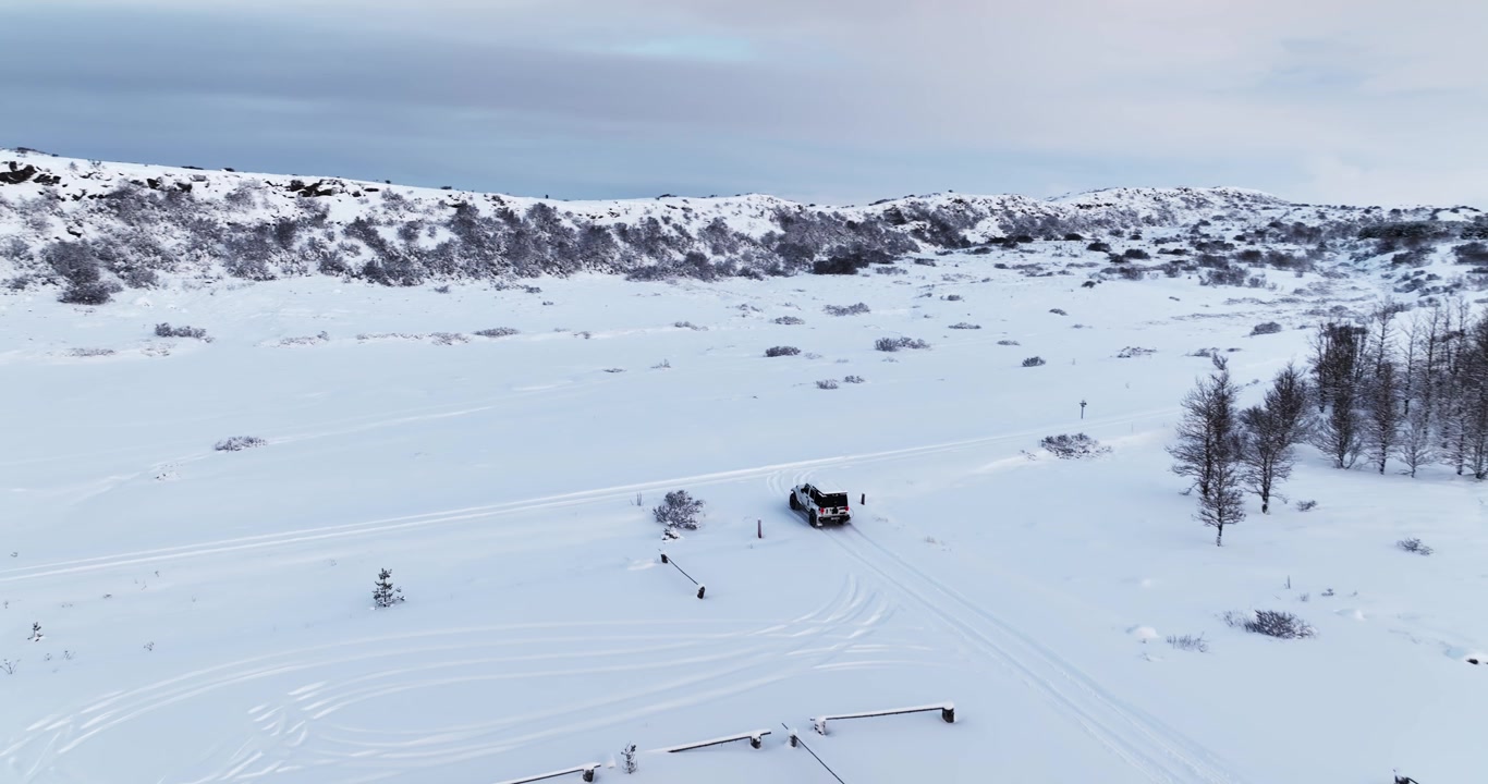 Fuoristrada bianco 4x4 che esce da un'area di parcheggio innevata a Heiðmörk, Islanda, con tracce di pneumatici, barriere di parcheggio, basse colline invernali e terreno ghiacciato