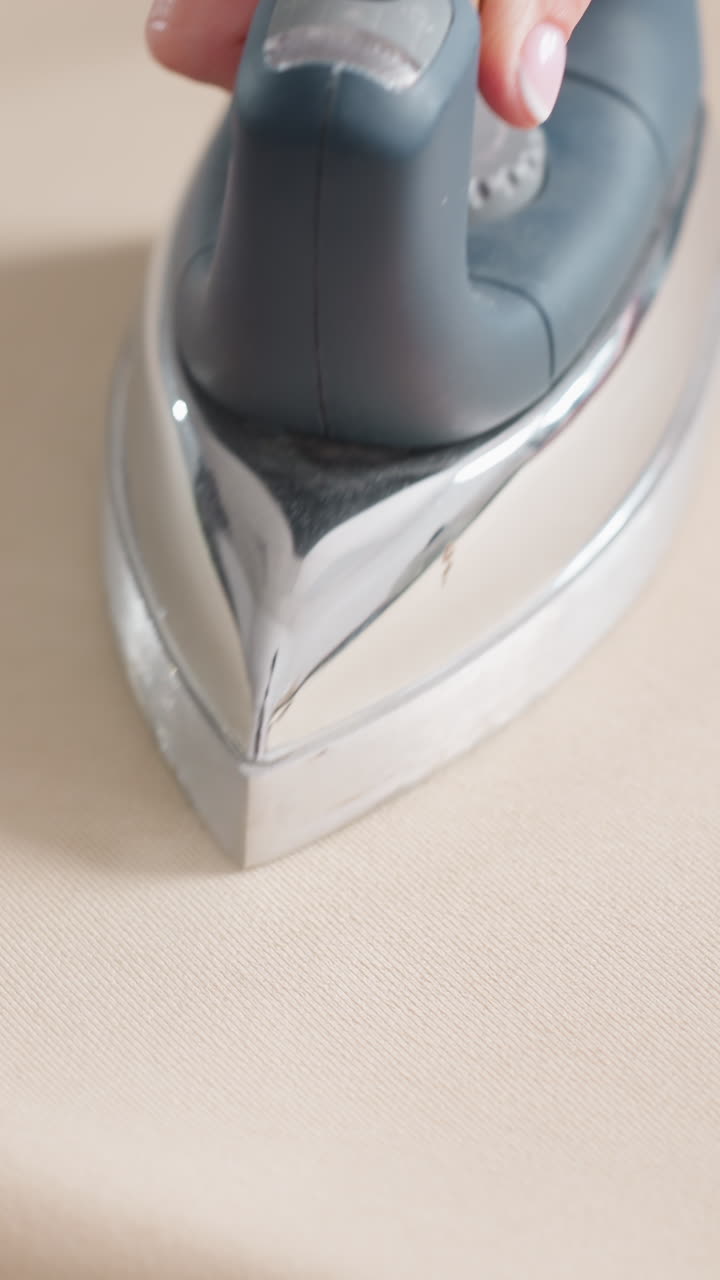 다림질로 천을 다듬는 모습이에요. 아틀리에 분위기 속에서 꾸준히 다림질하여 주름 없는 표면을 만들고, 베이지색 직물 위에 다리미 밑판이 매끄럽게 반사되는 모습이 보입니다. 깔끔한 마무리를 위한 차분하고 반복적인 동작이에요.