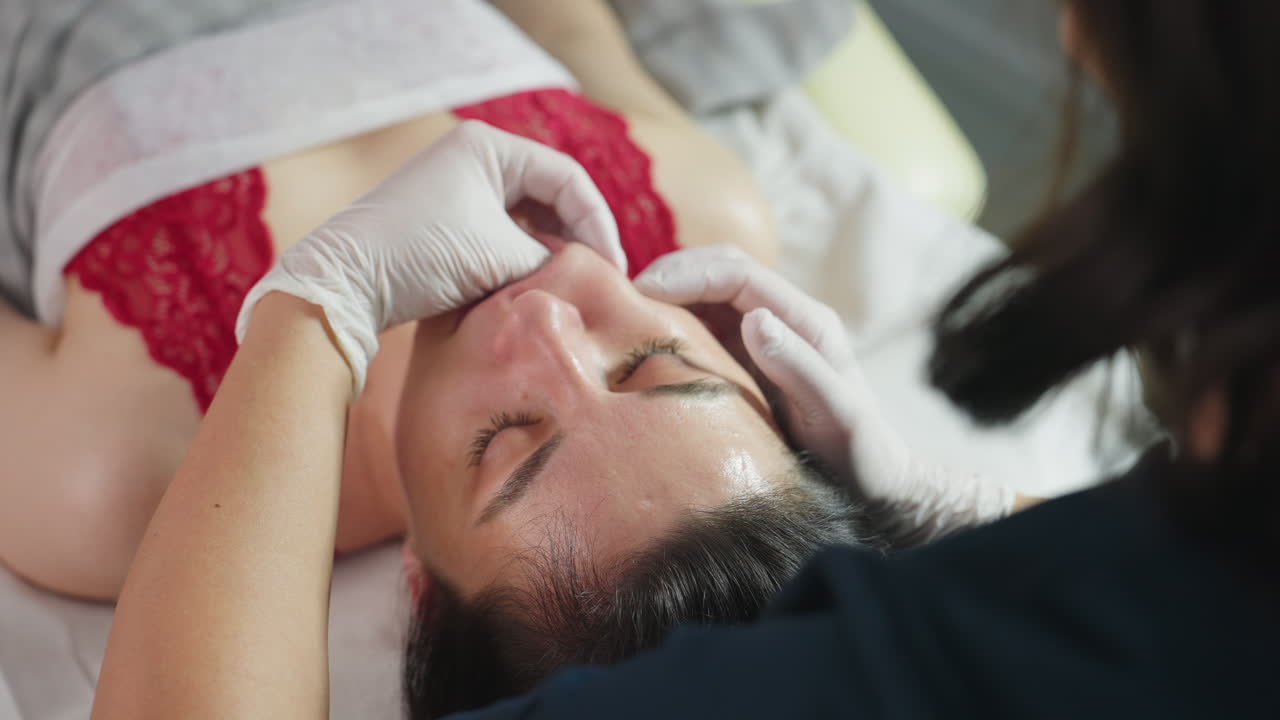 Female model on red sleeveless top closes eyes gently as bodywork expert wearing disposable gloves dips fingernail inside mouth and uses hand to massage cheeks in clinic with skin care bottles nearby