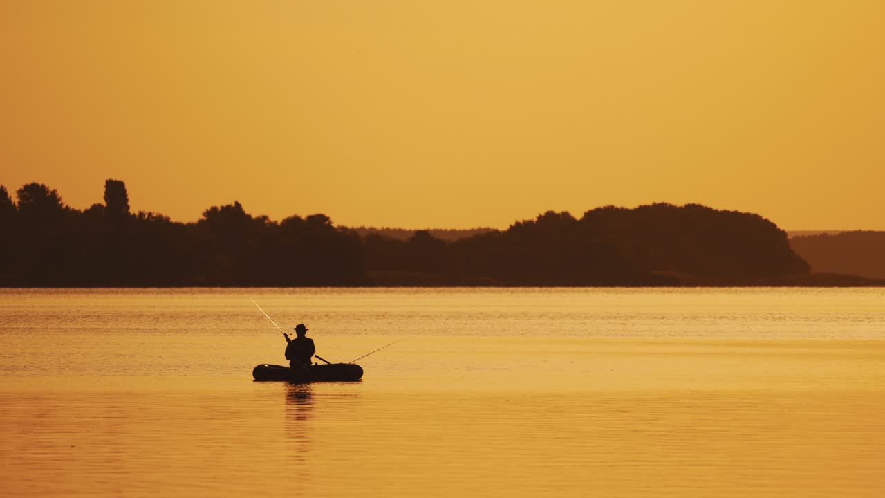 Amazing view of the evening sunset and a man in a boat fishing. Silhouette of a fisherman sitting in a row boat and catching fish on the golden evening background over the river surface.