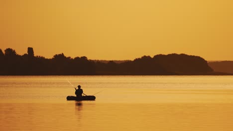 amazing view of the evening sunset and a man in a boat fishing. silhouette of a fisherman sitting in a row boat and catching fish on the golden evening background over the river surface.