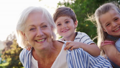 Portrait-Of-Smiling-Grandparents-Carrying-Grandchildren-Outdoors-In-Summer-Park