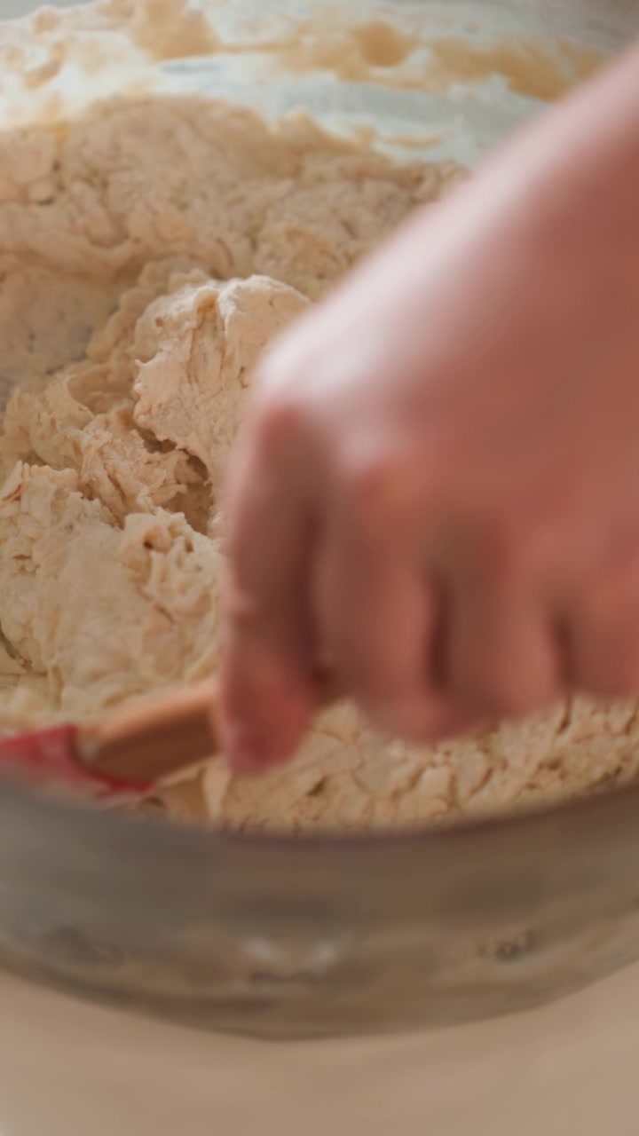 Close-up top-down scene of hand using wooden-handled spatula to mix shaggy dough in stainless steel bowl, breaking dry flour pockets, scraping sides, and folding ingredients together