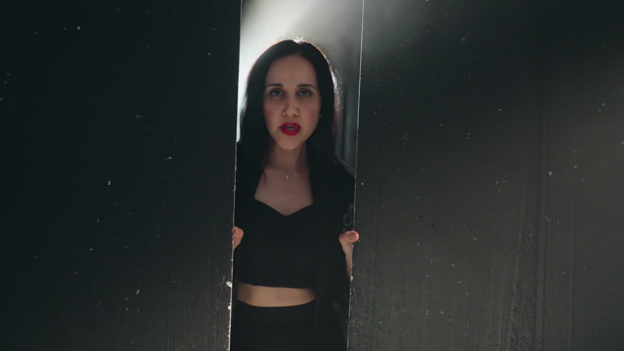 Elegant woman in black outfit and red lipstick partially visible behind a black wood wall, singing in soft light as camera zooms in and out
