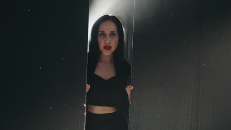 elegant woman in black outfit and red lipstick partially visible behind a black wood wall, singing in soft light as camera zooms in and out