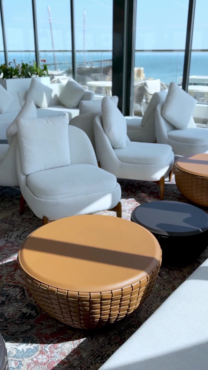 Vertical view of an empty modern hotel lounge with white chairs and woven tables. Bright daylight shines through large windows offering a scenic ocean view in this relaxing seaside resort interior.